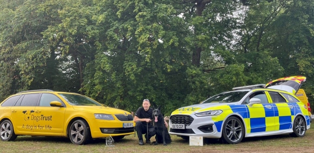 A police dog, Luther, poses with his handler in front of a police car and a Dogs Trust car